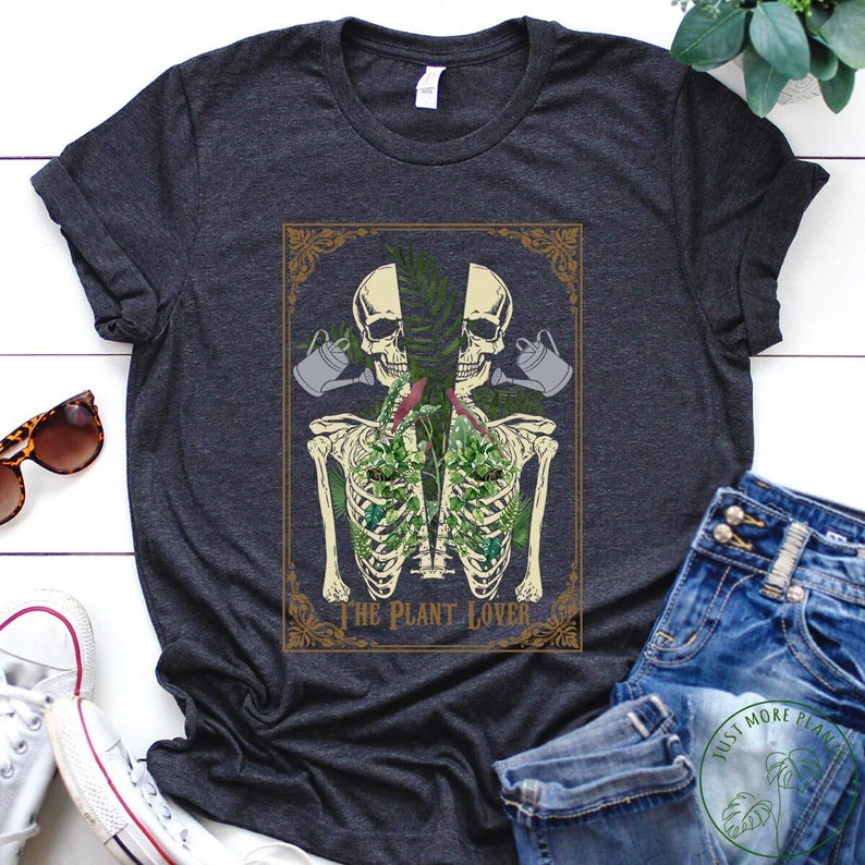 Peut inclure: T-shirt gris fonc&eacute; avec une illustration botanique. Le motif comprend un squelette avec des plantes poussant dans sa poitrine, des cr&acirc;nes et des arrosoirs. Le texte "THE PLANT LOVER" est au bas du motif.