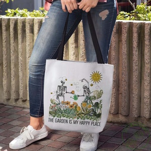 May include: A white tote bag with a design of two skeletons gardening. The skeletons are holding a watering can and a cup of tea. The text on the bag reads "The Garden is My Happy Place".