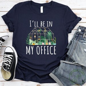 May include: A navy blue t-shirt with white text that reads "I'll be in my office". The text is above a graphic of a greenhouse filled with plants.