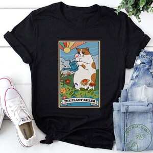 May include: Black t-shirt with a graphic of a cat holding a watering can in a garden with the text "The Plant Killer".