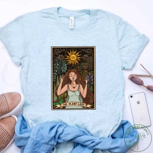 Pode incluir: Camiseta azul claro com um design de carta de tar&ocirc; com uma mulher cercada por plantas e o texto "The Plant Lady".