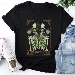 Peut inclure: T-shirt noir avec un motif graphique d'un torse de squelette rempli de plantes, deux t&ecirc;tes de mort et des arrosoirs. Le texte "THE PLANT LOVER" est en dessous. Des baskets blanches et un jean bleu sont visibles.