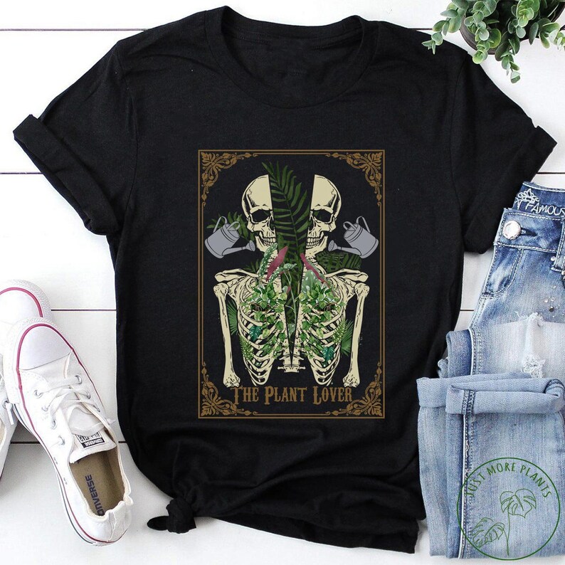 Peut inclure: T-shirt noir avec un motif graphique d'un torse de squelette rempli de plantes, deux t&ecirc;tes de mort et des arrosoirs. Le texte "THE PLANT LOVER" est en dessous. Des baskets blanches et un jean bleu sont visibles.