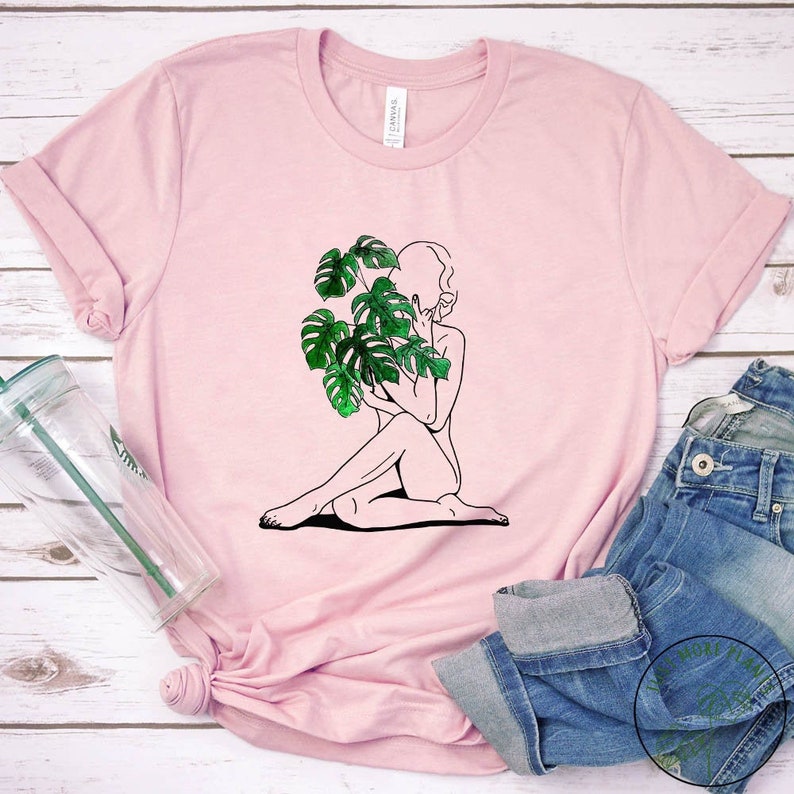 May include: A pink t-shirt featuring a black line drawing of a person sitting with a large green monstera plant in front of them.