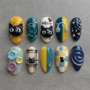 Peut inclure: Un ensemble de dix faux ongles présentant divers motifs, dont des visages de chats noirs, des tourbillons bleus, un ongle blanc avec le texte MEOW et une coccinelle, des écouteurs, des étoiles, des boutons, des carreaux et des damiers.
