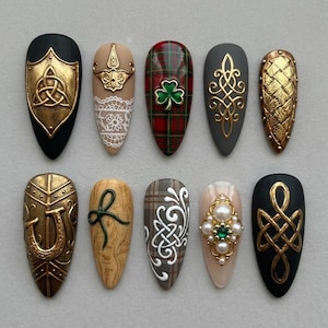 May include: Ten almond-shaped artificial nails with various designs. Features include gold Celtic knots, shields, and a horseshoe. Other nails have plaid, lace, and pearl embellishments. The colour palette includes black, gold, beige, red, and green.
