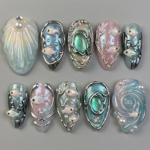 Peut inclure: Collection de dix créations d'art d'ongles sculptées et iridescentes. Chaque ongle présente un design aquatique unique, avec des poissons, des bulles et des perles. La palette de couleurs comprend des nuances de bleu, de rose et d'argent, créant un effet sous-marin chatoyant.