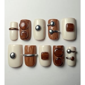 May include: A set of ten artificial nails with a variety of designs. Some nails are off-white with silver or brown accents. Other nails feature a wood grain pattern with silver or brown embellishments. The nails are arranged on a white surface.