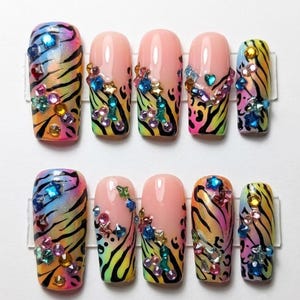 Peut inclure: Un ensemble de dix faux ongles avec un dégradé de rose, jaune et bleu orné de rayures de tigre noires. Chaque ongle est décoré de divers strass colorés aux formes variées, notamment des étoiles, des cœurs et des cercles disposés en groupes.