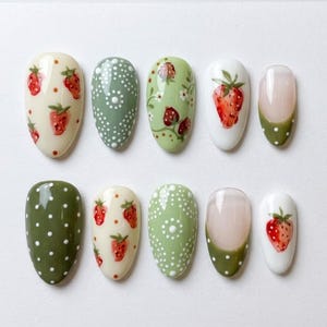 Peut inclure: Un ensemble de dix faux ongles en forme d'amande présentant divers motifs, notamment des fraises rouges sur fond crème et blanc, du vert sauge à pois blancs, des motifs floraux et des pointes françaises vertes à pois blancs sur base claire.