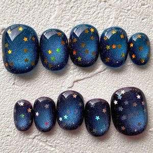 Peut inclure: Ensemble de faux ongles ovales. Ils présentent une base bleu foncé pailletée, ornée d'étoiles dorées et argentées. L'arrangement en deux rangées évoque un thème céleste, idéal pour la décoration des ongles.
