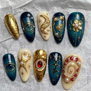 May include: A collection of ten artificial nails with intricate designs. The nails feature a variety of colors, including gold, teal, and cream. Some nails have gold embellishments, pearls, and celestial-themed designs like moons, stars, and eyes. The nails are oval-shaped.