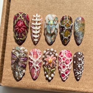 Peut inclure: Une collection de dix faux ongles aux motifs complexes. Chaque ongle présente des œuvres d'art détaillées, notamment des motifs floraux, des motifs d'insectes et des motifs abstraits dans diverses couleurs comme le rouge, le blanc, le marron et l'or. Les ongles sont disposés sur une surface brun clair.