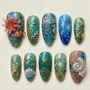 Peut inclure: Dix faux ongles sur le thème marin. Chaque ongle présente des motifs détaillés de créatures marines, dont des poissons, méduses, hippocampes et coraux, dans des tons de bleu, vert et or. Certains ongles ont des éléments en 3D.