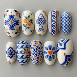 Peut inclure: Dix faux ongles ovales blancs avec des motifs peints à la main. Les motifs comprennent des motifs floraux bleus, des agrumes oranges, un poisson bleu et des motifs géométriques. Des accents de feuilles d'or ornent certains ongles. Thème : esthétique méditerranéenne.
