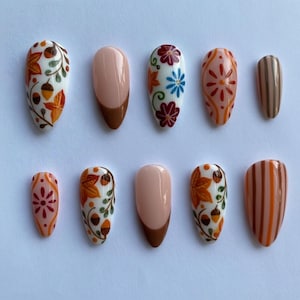 Peut inclure: Un ensemble de dix faux ongles avec divers motifs sur le thème de l'automne. Les ongles présentent des motifs de feuilles, de glands et de motifs floraux dans des tons orange, marron et blanc. Certains ongles ont un design à la française avec une base marron.