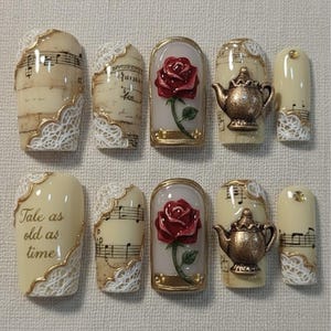 Peut inclure: Dix faux ongles décorés sur le thème de La Belle et la Bête. Les motifs incluent des partitions musicales, une rose rouge, une théière et la phrase "Tale as old as time". Les ongles présentent des accents de dentelle dorée et blanche.