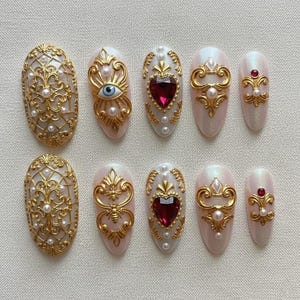 May include: A set of ten almond-shaped press-on nails. Each nail features a pearlescent base with ornate gold embellishments, including filigree, pearls, and heart-shaped gems. Some nails have an eye design or a bee.