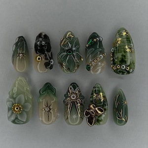 May include: A collection of ten artificial nails in diverse designs. The nails showcase green, gold, and beige hues, with detailed floral, geometric, and abstract patterns. Some nails have gold embellishments and gems.