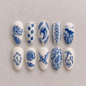 May include: A set of ten white oval-shaped fingernails, each adorned with blue dinosaur-themed illustrations. Designs include a T-Rex skull, footprints, a pterodactyl, bones, and a fossil. The nails are arranged on a white surface.
