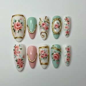 May include: A set of ten artificial nails with a floral design. The nails feature a variety of shapes and colors, including white, pink, and mint green, with gold accents. Each nail is decorated with hand-painted roses.