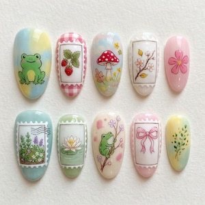 Peut inclure: Dix faux ongles décorés de motifs variés. Les motifs incluent une grenouille, des fraises, un champignon, des fleurs et un nœud. Les ongles sont peints dans des couleurs pastel, notamment rose, jaune, bleu et vert.