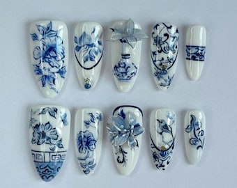 May include: A set of ten white artificial nails with blue floral designs, reminiscent of porcelain. Some nails feature 3D flower accents and small jewels. The designs include floral patterns, vases, and bird motifs, creating a cohesive aesthetic.