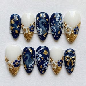 May include: A set of ten artificial nails with a navy blue, white, and gold design. The nails feature floral patterns, gold leaf accents, and a swan illustration. Some nails have a gold bow design.