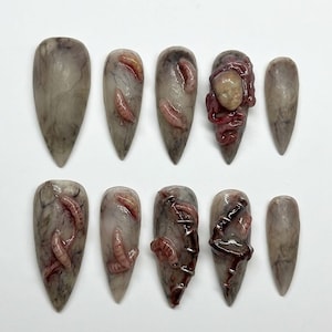 Peut inclure: Un ensemble de dix faux ongles sur le thème macabre. Les ongles sont principalement gris avec un effet marbré et présentent des détails réalistes et grotesques tels que des vers et un petit visage, créant une esthétique troublante.
