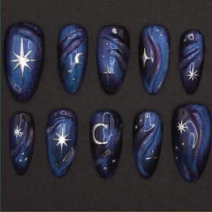 May include: A set of ten press-on nails with a dark blue and purple galaxy design. Each nail features a different celestial element, including stars and crescent moons, in silver. The nails have a glossy finish.