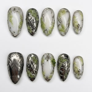 Peut inclure: Un ensemble de dix faux ongles avec une finition métallique argentée. Chaque ongle présente un design unique, avec des accents blancs et verts et des motifs complexes. Les ongles sont de forme ovale et disposés en deux rangées de cinq.