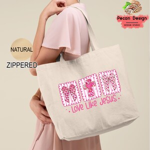 May include: A natural canvas tote bag with a zippered closure. The bag features a pink and white design with bows, a cross, and the text "Love Like Jesus". The bag is being held by a person.