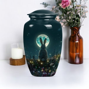 Puede incluir: Una urna de cerámica azul oscuro con una escena de luna llena que presenta un conejo mirando hacia la luna. La urna está decorada con un diseño caprichoso de flores, luciérnagas y estrellas.
