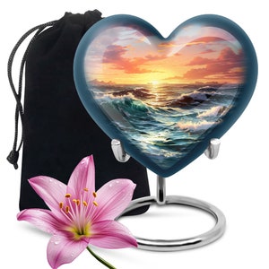 May include: A heart-shaped glass keepsake urn with a sunset over the ocean image. The urn is on a silver stand and comes with a black velvet bag.