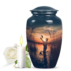 May include: A dark blue cremation urn featuring a sunset scene of a person fishing in a lake. The urn has a lid and a white candle is in front of it.