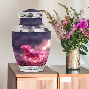 May include: A silver cremation urn featuring a pink unicorn galloping through a stormy sky. The urn is decorated with an intricate design around the top and bottom.
