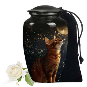 Puede incluir: Urna de cerámica negra para mascotas con un gato atigrado marrón mirando hacia la luna y las estrellas. La urna viene con una bolsa de terciopelo negro.