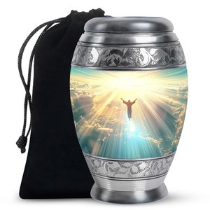May include: Silver metal urn featuring a religious image of a person with outstretched arms in front of a bright light and clouds. The urn has a decorative floral design around the top and bottom. It comes with a black velvet bag.