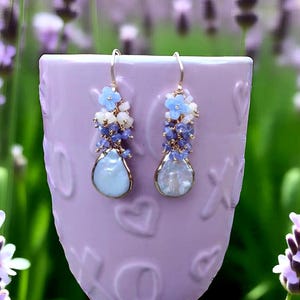 May include: A pair of gold-toned earrings with a floral design. Each earring features a cluster of light blue flowers, white beads, and blue gemstones, with a teardrop-shaped, pale blue pendant. The earrings are displayed against a lavender background.