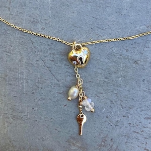 May include: A gold-tone necklace featuring a heart-shaped pendant with a central keyhole design and two small inset stones. A delicate chain drop hangs from the heart, adorned with a white pearl, two translucent beads, and a small gold-tone key charm.