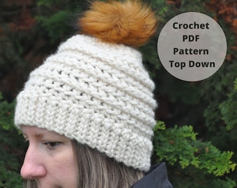 May include: A white crocheted beanie with a brown faux fur pom pom. The beanie is being worn by a person. The text "Crochet PDF Pattern Top Down" is visible in the image.