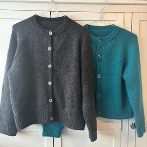Peut inclure: Deux cardigans tricotés suspendus à un cintre métallique. L'un est gris anthracite, l'autre est turquoise. Les deux ont un col rond, des manches longues et des fermetures à boutons. Les cardigans sont faits d'un fil doux et texturé.