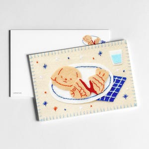 Può includere: Una cartolina in feltro stravagante con un cane dei cartoni animati e cibo su un piatto beige. Include un tovagliolo a scacchi blu e bianco e un bicchiere d'acqua. Lo sfondo è beige chiaro con dettagli di stelle blu e arancioni.