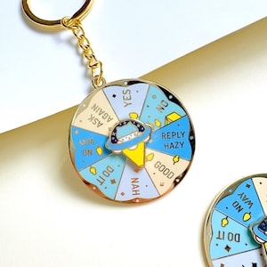 May include: A gold metal keyring with a circular spinner design. The spinner is in shades of blue and white, with phrases such as "Yes", "No", and "Ask Again". A small, cartoon spaceship is in the centre of the spinner.