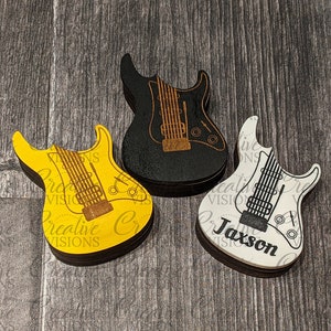 May include: Three wooden guitar-shaped ornaments, one yellow, one white, and one black. The white guitar has the name "Jaxson" written on it.