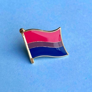 May include: A gold-coloured pin featuring a bisexual pride flag design. The flag has three horizontal stripes: pink, purple, and blue.