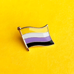 May include: A yellow, white, purple and black enamel pin in the shape of a flag. The pin is on a yellow background.