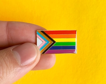 May include: A rectangular enamel pin featuring the Progress Pride flag design. The pin has a rainbow stripe design with additional stripes in pink, light blue, brown, black, and white. The pin is held against a yellow background.