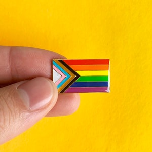 May include: A rectangular enamel pin displaying the Progress Pride flag. The pin features a rainbow design with additional stripes in pink, light blue, brown, black, and white. The pin is set against a yellow background.