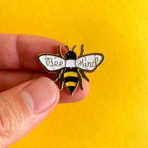 Op de afbeelding: Gouden bij-emaille pin met zwarte en gele strepen en witte vleugels. Op de vleugels staat "Bee Kind".